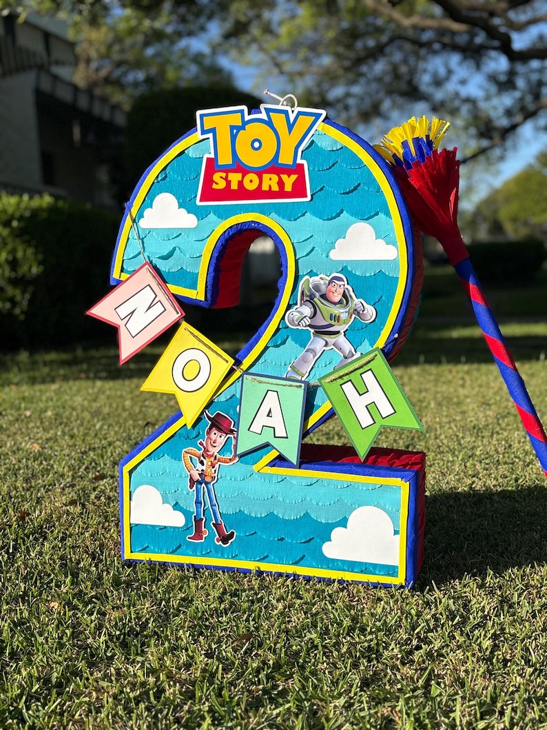 May include: A number two shaped pi&ntilde;ata featuring a blue sky background with white clouds, the Toy Story logo, and character cutouts of Buzz Lightyear and Woody. A banner hanging across the front displays the name NOAH in individual flag letters.