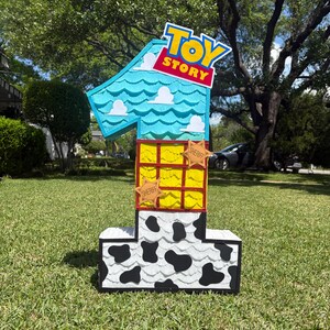 May include: A number one shaped pi&ntilde;ata featuring a Toy Story theme. The top section is light blue with white clouds, the middle is yellow with a red grid and two sheriff star badges, and the base is white with black cow print patterns.
