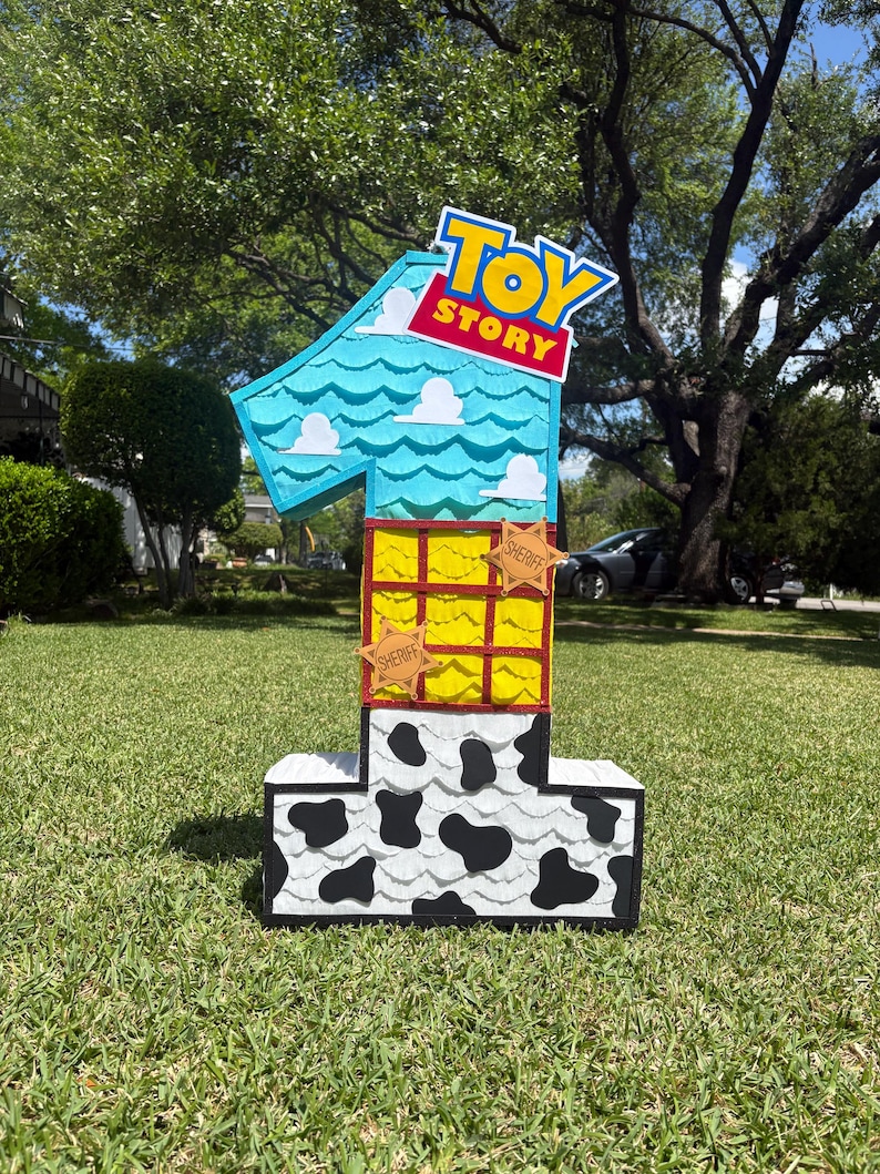 May include: A number one shaped pi&ntilde;ata featuring a Toy Story theme. The top section is light blue with white clouds, the middle is yellow with a red grid and two sheriff star badges, and the base is white with black cow print patterns.