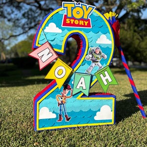 May include: A number two shaped pinata featuring a blue background with a scalloped pattern, white clouds, and the Toy Story logo. It includes cutouts of Buzz Lightyear and Woody, along with a banner spelling the name NOAH in colorful flags.