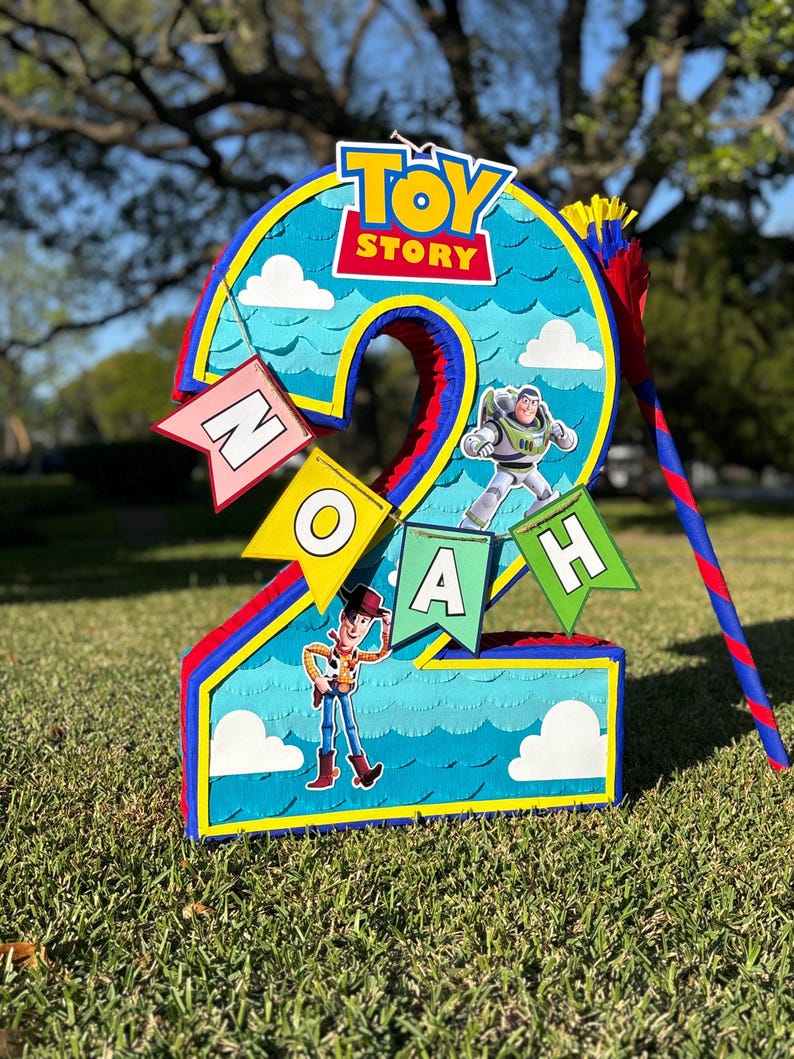 May include: A number two shaped pinata featuring a blue background with a scalloped pattern, white clouds, and the Toy Story logo. It includes cutouts of Buzz Lightyear and Woody, along with a banner spelling the name NOAH in colorful flags.