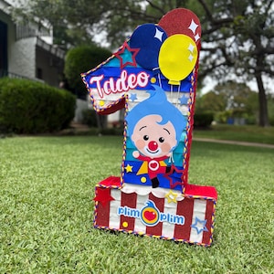 Puede incluir: Una pi&ntilde;ata con forma de n&uacute;mero uno decorada con rayas rojas, blancas y azules, que presenta un personaje de dibujos animados con pelo azul y nariz roja, el nombre Tadeo en letra blanca y el logo de Plim Plim con un coraz&oacute;n y globos.