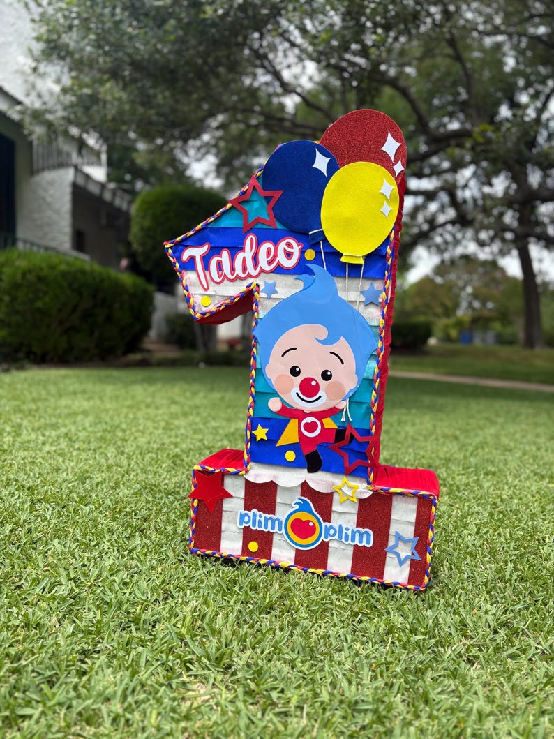 Puede incluir: Una pi&ntilde;ata con forma de n&uacute;mero uno decorada con rayas rojas, blancas y azules, que presenta un personaje de dibujos animados con pelo azul y nariz roja, el nombre Tadeo en letra blanca y el logo de Plim Plim con un coraz&oacute;n y globos.