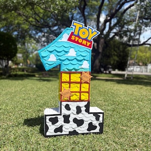 May include: A number one shaped pinata featuring a Toy Story theme. The top section is light blue with white clouds, the middle is yellow with a red grid, and the bottom is white with black cow print spots. Two brown sheriff star badges are attached.
