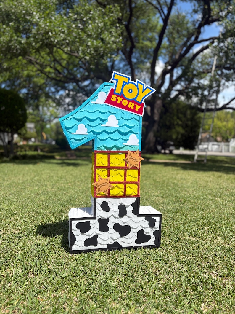 May include: A number one shaped pinata featuring a Toy Story theme. The top section is light blue with white clouds, the middle is yellow with a red grid, and the bottom is white with black cow print spots. Two brown sheriff star badges are attached.
