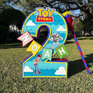 May include: A number two shaped pinata featuring a blue sky background with white clouds, the Toy Story logo, and images of Buzz Lightyear and Woody. A banner with the name NOAH is attached, and a red and blue striped stick is included.