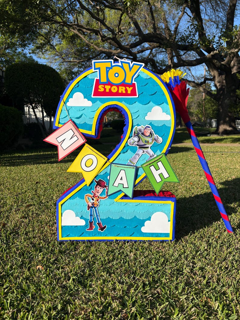 May include: A number two shaped pinata featuring a blue sky background with white clouds, the Toy Story logo, and images of Buzz Lightyear and Woody. A banner with the name NOAH is attached, and a red and blue striped stick is included.