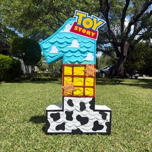 May include: A number one shaped pinata featuring a Toy Story theme. The top section is light blue with white clouds, the middle is yellow with a red grid, and the base is white with black cow print spots. Two tan sheriff star badges are attached.