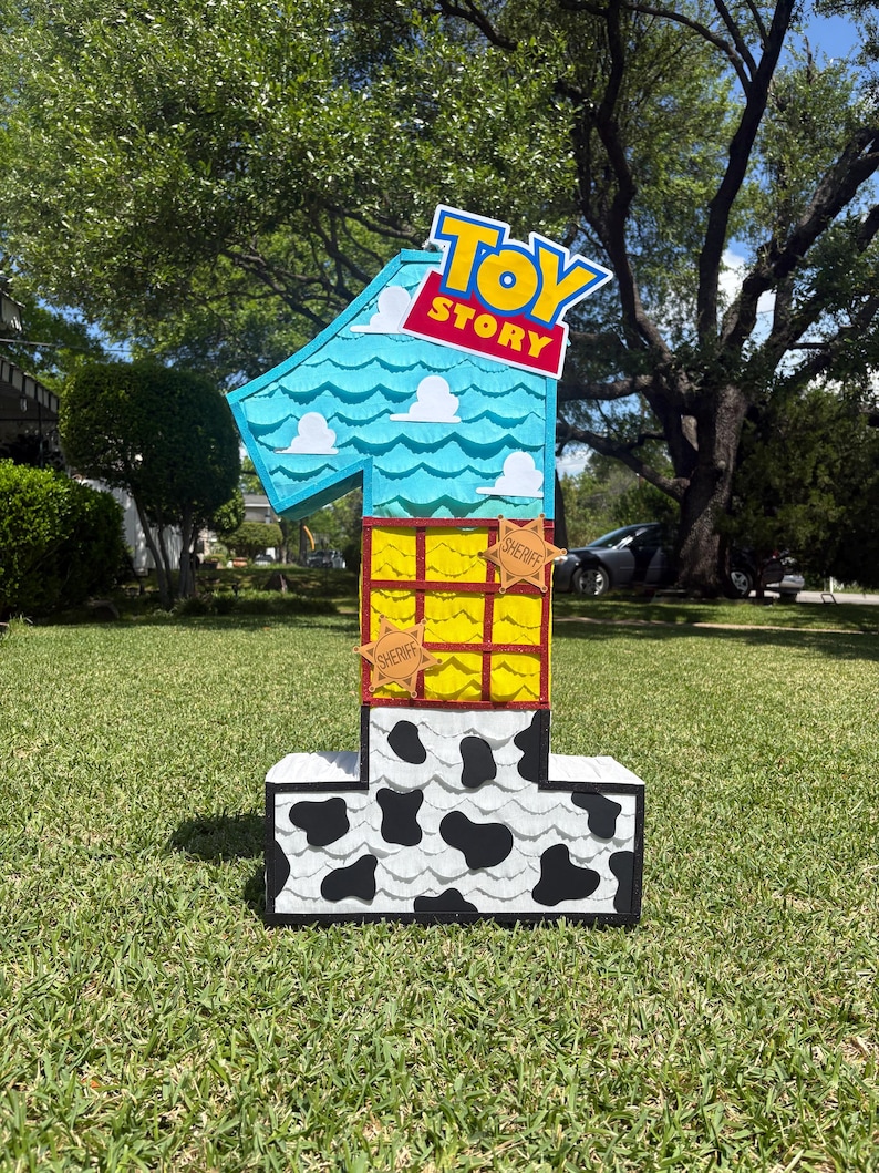 May include: A number one shaped pinata featuring a Toy Story theme. The top section is light blue with white clouds, the middle is yellow with a red grid, and the base is white with black cow print spots. Two tan sheriff star badges are attached.