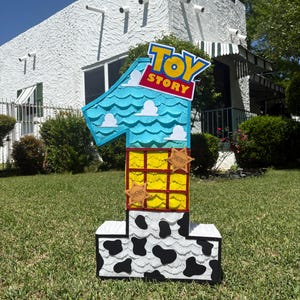 May include: A large number one shaped pinata featuring a Toy Story theme. The top section is light blue with white clouds, the middle is yellow with a red grid pattern and two sheriff badges, and the base is white with black cow print spots.