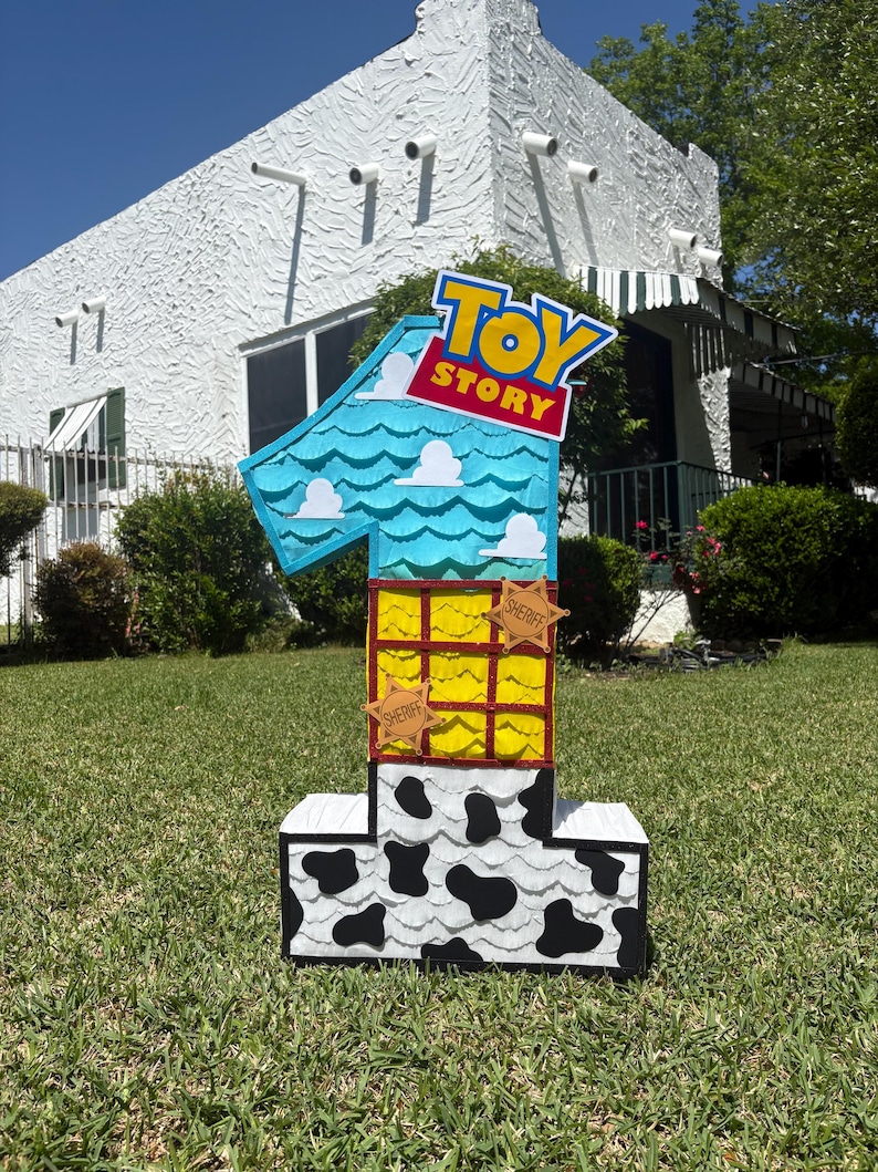 May include: A large number one shaped pinata featuring a Toy Story theme. The top section is light blue with white clouds, the middle is yellow with a red grid pattern and two sheriff badges, and the base is white with black cow print spots.