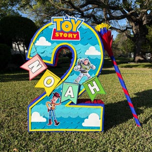 May include: A number two shaped pinata featuring a blue sky background with white clouds, the Toy Story logo, and character cutouts of Woody and Buzz Lightyear. A banner spells out the name Noah, and a matching blue and red striped stick is included.