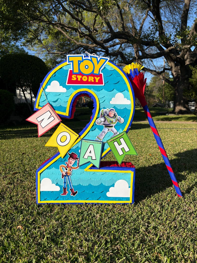 May include: A number two shaped pinata featuring a blue sky background with white clouds, the Toy Story logo, and character cutouts of Woody and Buzz Lightyear. A banner spells out the name Noah, and a matching blue and red striped stick is included.