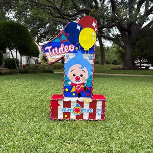 Puede incluir: Una pi&ntilde;ata con forma de n&uacute;mero uno que presenta un personaje de dibujos animados azul con nariz roja, el nombre Tadeo en letra blanca y el texto plim plim. El dise&ntilde;o incluye rayas rojas, blancas y azules, globos amarillos y rojos.