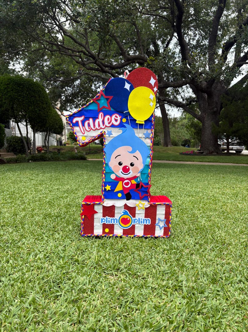 Puede incluir: Una pi&ntilde;ata con forma de n&uacute;mero uno que presenta un personaje de dibujos animados azul con nariz roja, el nombre Tadeo en letra blanca y el texto plim plim. El dise&ntilde;o incluye rayas rojas, blancas y azules, globos amarillos y rojos.