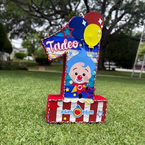 Puede incluir: Una pi&ntilde;ata con forma de n&uacute;mero uno que presenta un personaje de dibujos animados azul con nariz roja, el nombre Tadeo en letra blanca y el logotipo de Plim Plim. El dise&ntilde;o incluye rayas rojas y blancas, detalles brillantes y estrellas y globos.
