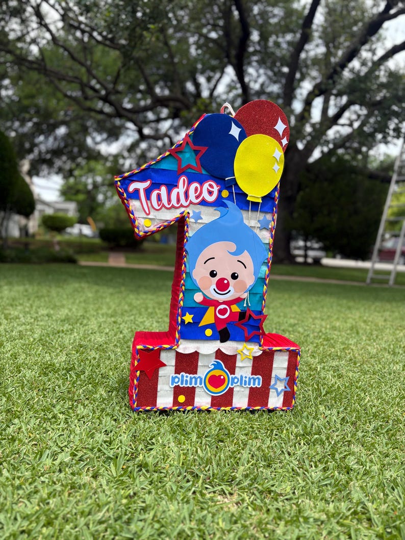 Puede incluir: Una pi&ntilde;ata con forma de n&uacute;mero uno que presenta un personaje de dibujos animados azul con nariz roja, el nombre Tadeo en letra blanca y el logotipo de Plim Plim. El dise&ntilde;o incluye rayas rojas y blancas, detalles brillantes y estrellas y globos.