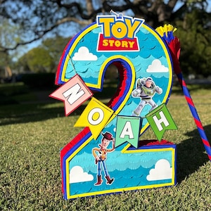 May include: A number two shaped pinata featuring a blue sky background with white clouds, the Toy Story logo, and character cutouts of Woody and Buzz Lightyear. A banner with the name NOAH is attached to the front in colorful pennant flags.