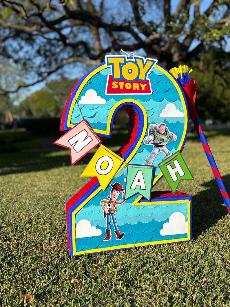 May include: A number two shaped pinata featuring a blue sky background with white clouds, the Toy Story logo, and character cutouts of Woody and Buzz Lightyear. A banner with the name NOAH is attached to the front in colorful pennant flags.