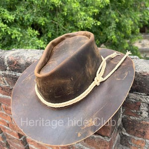 May include: A brown leather hat with a distressed finish, featuring a wide brim and a cream-colored braided rope band tied in a knot around the crown. The text "Heritage hide Club" is printed in white across the lower portion of the brim.