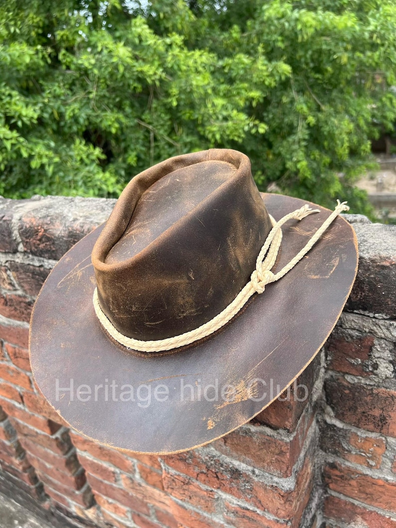 May include: A brown leather hat with a distressed finish, featuring a wide brim and a cream-colored braided rope band tied in a knot around the crown. The text "Heritage hide Club" is printed in white across the lower portion of the brim.