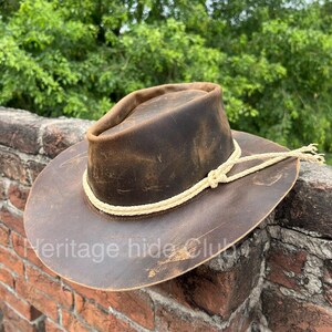May include: A dark brown distressed leather hat with a wide brim and a pinched crown. A cream-colored braided rope band encircles the base of the crown, tied in a simple knot on the side. The text Heritage hide Club is visible on the brim.