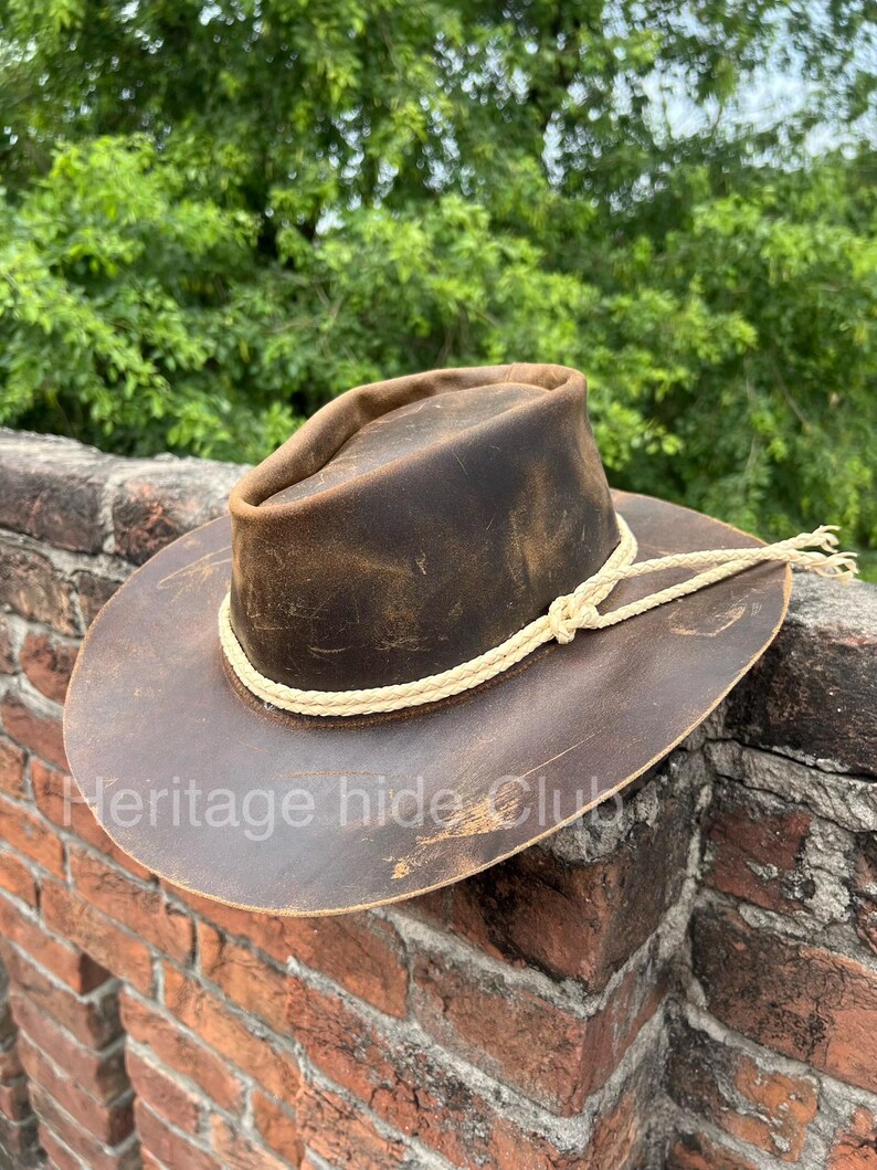 May include: A dark brown distressed leather hat with a wide brim and a pinched crown. A cream-colored braided rope band encircles the base of the crown, tied in a simple knot on the side. The text Heritage hide Club is visible on the brim.