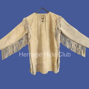 May include: A tan suede shirt with long sleeves and fringe detailing along the sleeves and hem. The shirt has a laced neckline and the text "Heritage Hide Club" is printed on the front.