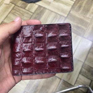 May include: A burgundy leather wallet with a crocodile skin texture. The rectangular wallet has a black stitched border. The wallet is held in a person's hand.
