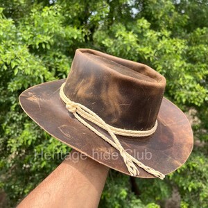 May include: A brown leather hat with a distressed finish and a wide brim. The hat features a decorative cream-colored braided cord wrapped around the base of the crown, tied with a knot on the side, with two loose braided ends hanging down.