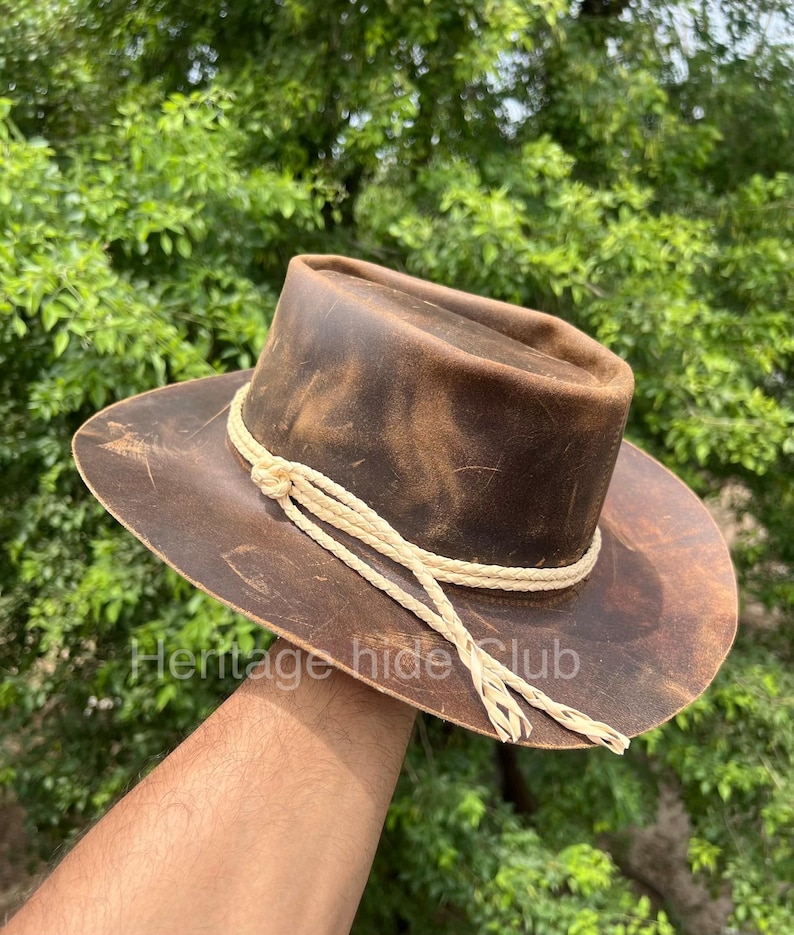 May include: A brown leather hat with a distressed finish and a wide brim. The hat features a decorative cream-colored braided cord wrapped around the base of the crown, tied with a knot on the side, with two loose braided ends hanging down.