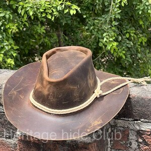 May include: A brown leather cowboy hat with a distressed finish and visible surface scratches. The hat features a pinched crown and a wide brim, accented by a braided cream-colored cord tied around the base of the crown with loose ends.