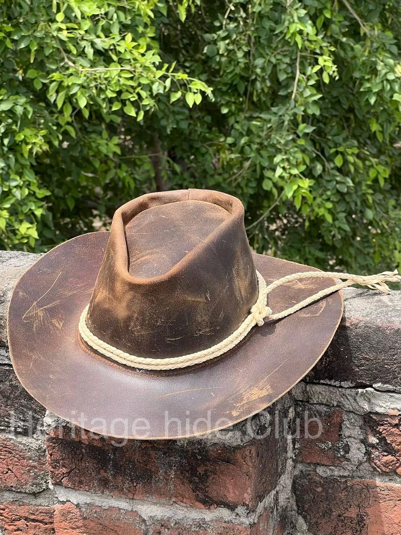 May include: A brown leather cowboy hat with a distressed finish and visible surface scratches. The hat features a pinched crown and a wide brim, accented by a braided cream-colored cord tied around the base of the crown with loose ends.