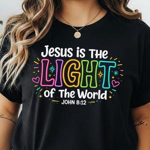 May include: A black short-sleeved t-shirt featuring the colorful text "Jesus is The LIGHT of The World" and "JOHN 8:12" in a neon-style font. The design includes small star and heart graphics in pink, yellow, blue, and green surrounding the text.