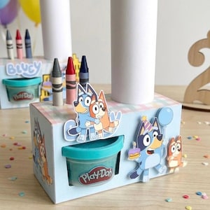 Puede incluir: Una caja azul claro con recortes de personajes de Bluey, recipientes de Play-Doh y crayones. Dos rollos de papel cilíndricos blancos están encima. La caja tiene un diseño azul claro y blanco. Globos y confeti en el fondo.