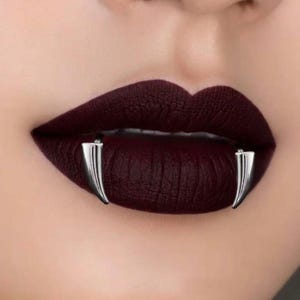 May include: A pair of silver-toned metal fang-shaped tooth covers positioned over the lower lip. The fangs have a smooth, polished finish and are worn against dark, matte-textured lipstick that covers the upper and lower lips.