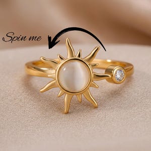 May include: A gold-colored ring featuring a sunburst design with a central, round, opalescent stone. The ring has a small, clear gemstone on the side. The text "Spin me" is above the ring with a curved arrow.