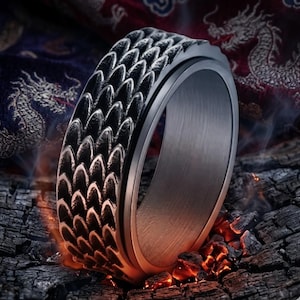 May include: A black and silver ring with a dragon scale pattern. The ring is set against a backdrop of burning embers and charred wood, with a hint of smoke and a dragon-themed textile in the background.