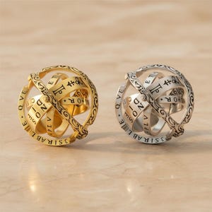 Könnte beinhalten: Zwei kugelförmige, bewegliche Ringe. Einer ist goldfarben, der andere silberfarben. Beide haben eingravierte Texte und komplizierte Designs. Die Ringe scheinen Schmuck oder dekorative Objekte zu sein, möglicherweise im Zusammenhang mit Astronomie oder historischen Themen.