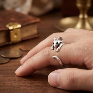 May include: A silver-colored ring in the shape of a mouse, with the mouse's head and paws wrapping around the finger. The mouse has black eyes and detailed ears. The ring's tail curves around the finger, creating a unique and whimsical design.