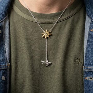 May include: A silver lariat necklace featuring a gold-toned sun charm at the centre point and a small silver human figure pendant dangling at the end of the chain. The necklace is worn over an olive green t-shirt and a blue denim jacket.