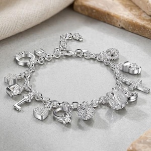 Puede incluir: Una pulsera de dijes de plata con múltiples colgantes, incluyendo una cruz, un corazón, una llave, una estrella y varios anillos, todos decorados con pequeñas piedras transparentes. La pulsera está formada por una serie de eslabones circulares.