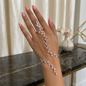 May include: A silver-toned hand chain featuring a delicate vine-like design adorned with marquise-cut clear crystals. The piece connects a ring worn on the middle finger to a bracelet encircling the wrist, creating a continuous floral motif.