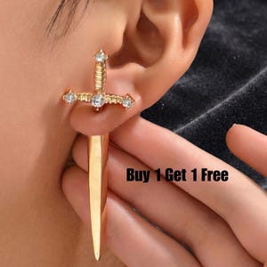 May include: Gold-colored sword-shaped earrings with clear crystal accents. The earrings are worn on an ear, with the sword's hilt and blade visible. The text "Buy 1 Get 1 Free" is in the lower right corner.