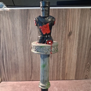 Könnte beinhalten: Eine schwarze Ritterbüste mit einem roten Emblem auf der Brust, ausgestattet mit einem Helm und fehlenden Gliedmaßen. Die Figur ist auf einer runden, verzierten Plattform montiert, die auf einer verwitterten, kannelierten Säule mit grünen und roten Farbakzenten steht.