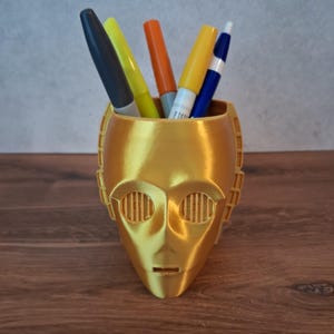 Könnte beinhalten: Ein goldfarbener, 3D-gedruckter Schreibtisch-Organizer in Form eines Roboterkopfes mit kreisförmigen, geschlitzten Augendetails. Das hohle Innere enthält fünf Schreibgeräte, darunter einen schwarzen Stift, einen gelben Textmarker, einen orangefarbenen Marker, einen gelben Marker sowie einen blau-weißen Stift.