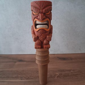Può includere: Un tappo per bottiglia in legno intagliato con un design di dio Tiki. Il Tiki ha un'espressione arrabbiata con accenti arancioni intorno agli occhi e alla bocca. Il tappo è di colore marrone rossiccio ed è posto su una superficie di legno.
