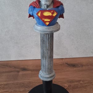 May include: A figurine featuring a skull-faced character with black hair, wearing a blue suit with a red cape and a yellow and red chest emblem. The character is mounted on a tall, fluted gray column that sits atop a circular black base.