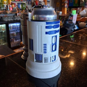 Könnte beinhalten: Ein weiß-blauer R2-D2-Kühler mit einer silbernen Dose im Inneren. Der Kühler hat blaue Akzente und Details, die dem ikonischen Star Wars-Droiden ähneln. Der Kühler ist zylindrisch und steht auf einem weißen Sockel.