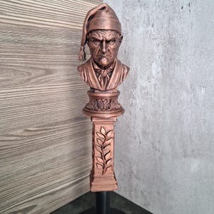 Może przedstawiać: Miedziany uchwyt do kranu z wyrzeźbionym popiersiem mężczyzny o surowym wyrazie twarzy w czapce nocnej. Popiersie spoczywa na kwadratowej podstawie ozdobionej pionowym reliefem z liści laurowych, wykończonej metaliczną patyną w kolorze brązu.